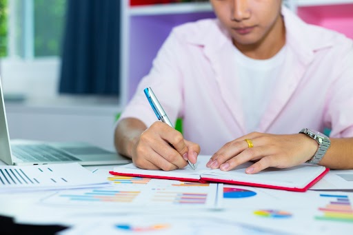 A person in a grey sweater holding a red pen and reviewing documents on a book pile, showcasing Scora’s expert help in academic proposal writing.